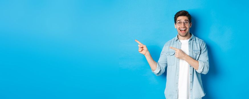 Excited handsome man in glasses pointing fingers left, looking happy while showing advertisement, demonstrating copy space on blue background.