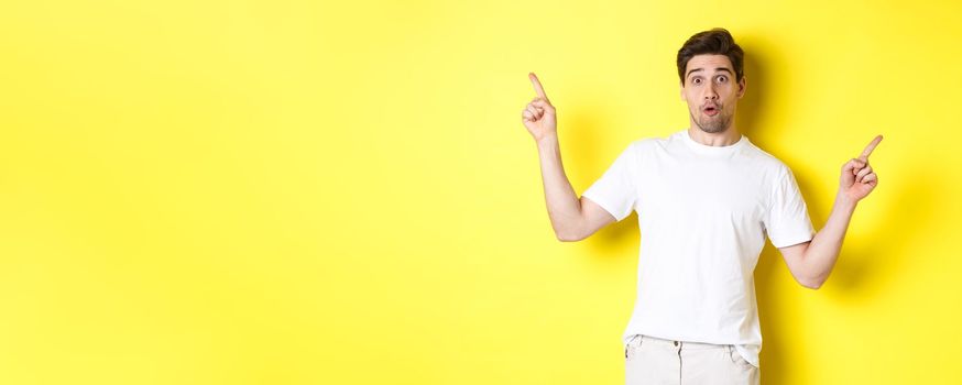 Handsome man pointing fingers sideways, showing two promos, standing over yellow background.