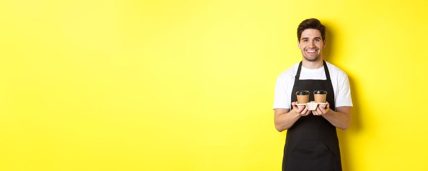 Handsome male barista serving takeaway coffee and smiling, bringing order, standing in black apron against yellow background.