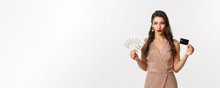 Shopping concept. Sassy woman in elegant dress showing credit card with money, looking pleased at camera, standing over white background. Copy space
