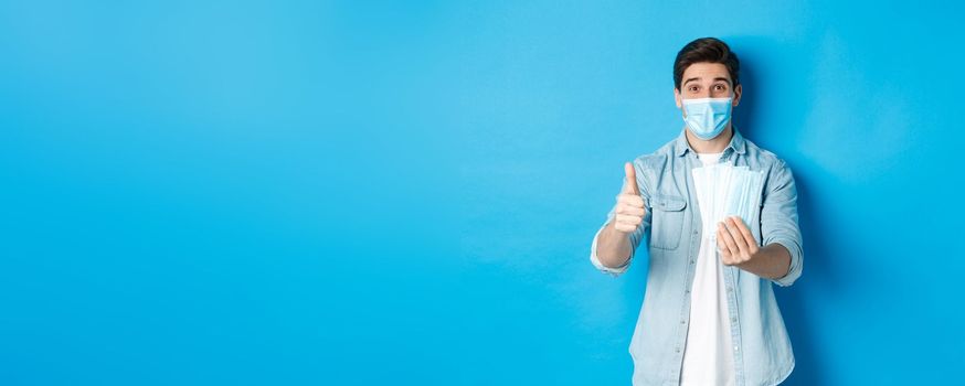 Concept of coronavirus, quarantine and social distancing. Young man showing medical masks and thumb up, standing against blue background.