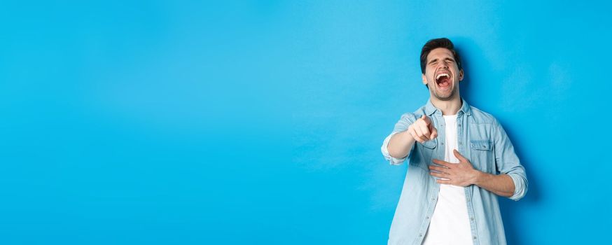 Adult man in casual outfit laughing out loud and pointing at you, looking at something funny, chuckling while standing against blue background.