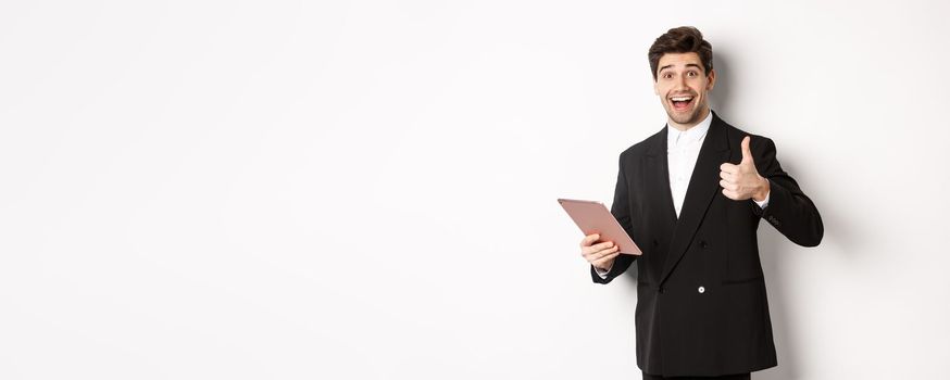 Portrait of handsome businessman in suit, showing thumb-up and making a compliment, praising something on digital tablet, standing over white background.