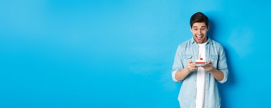 Excited young male model looking at birthday cake and smiling, making a wish and celebrating, standing against blue background.