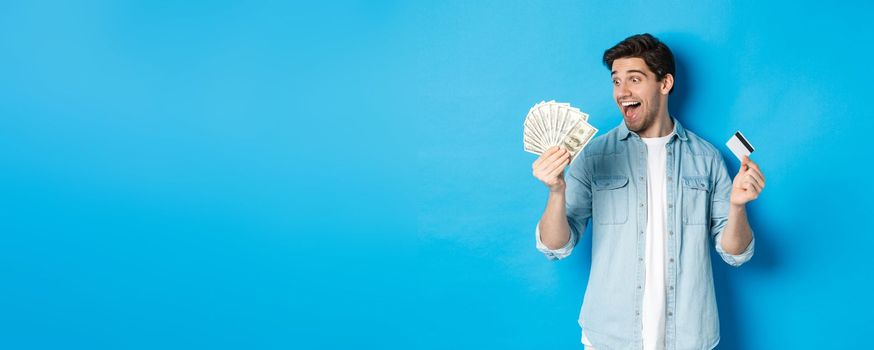 Amazed and happy man holding credit card, looking at money satisfied, standing over blue background.