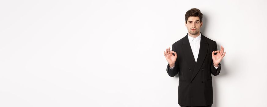 Concept of new year party, celebration and lifestyle. Portrait of confident good-looking man in black suit, showing okay sign and approve something, standing over white background.