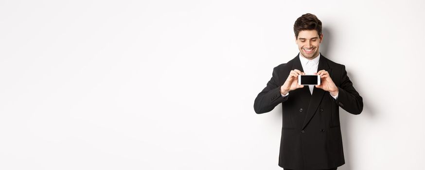 Portrait of handsome bearded man in black suit, showing you smartphone screen and smiling pleased, standing against white background.