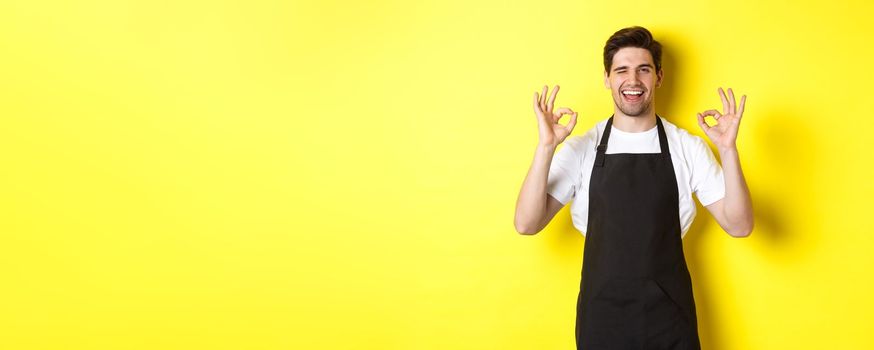 Young male barista in black apron showing OK signs and smiling, guarantee quality in coffee shop, standing over yellow background.