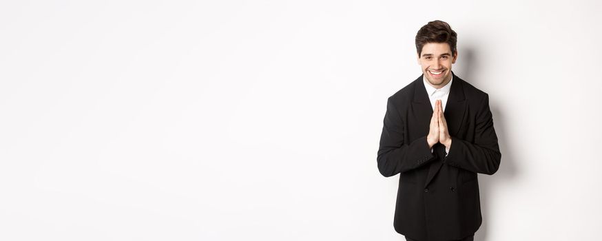 Portrait of handsome man in black suit, being grateful, saying thank you and bowing politely, smiling as holding hands together, expressing gratitude, standing over white background.