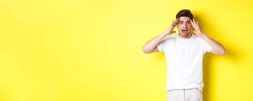 Frustrated guy looking shocked, panicking and holding hands on head, standing against yellow background.