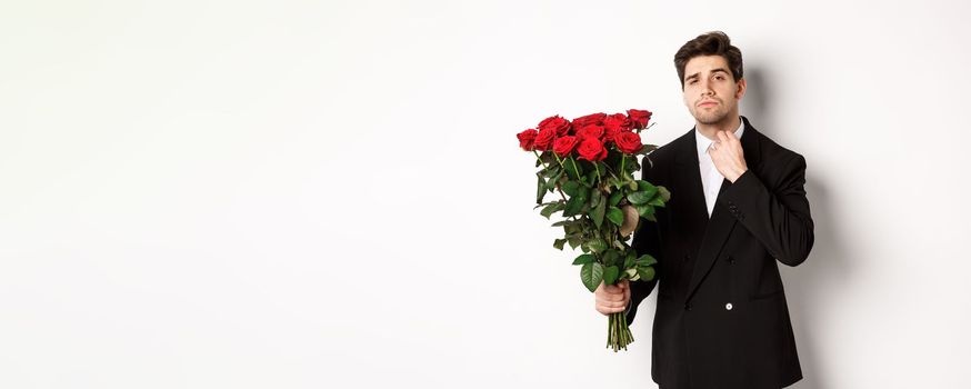 Image of elegant and sassy man in black suit, looking confident and holding bouquet of red roses, going on a romantic date, standing against white background.