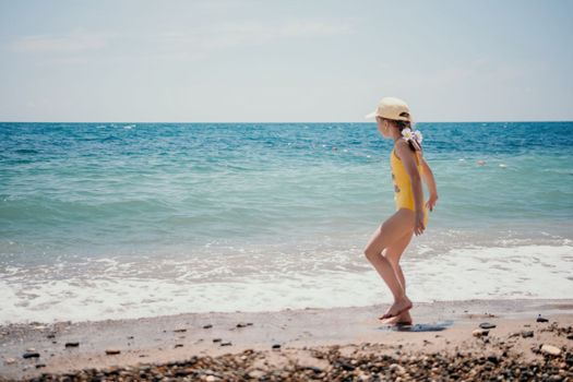 Cute little girl running along the seashore against a clear blue sea and rejoices in the rays of the summer sun. Beautiful girl in yellow swimsuit running and having fun on tropical beach