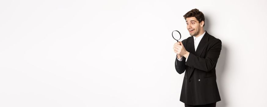 Image of handsome businessman in black suit, looking through magnifying glass, found something and smiling, standing over white background.