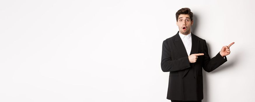 Portrait of surprised handsome man with beard, wearing black suit, open mouth and pointing fingers right at advertisement, standing over white background.