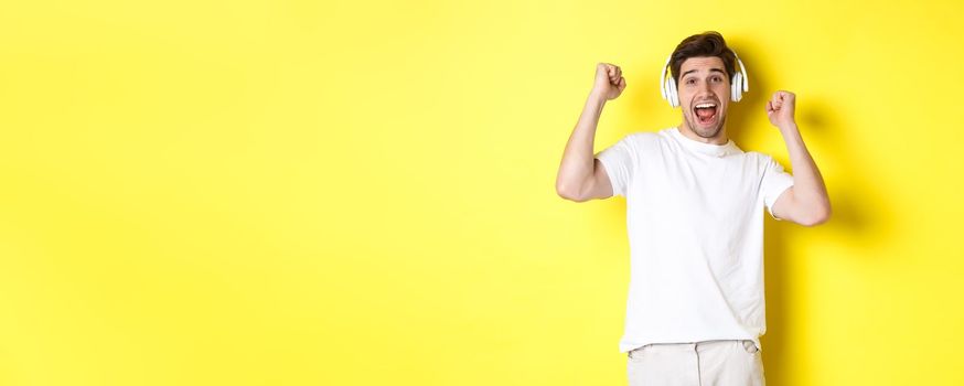 Excited handsome man dancing and singing along, listening music in headphones, standing over yellow background.