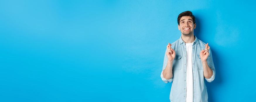 Lucky guy praying and making wish with crossed fingers, looking up with pleading face, standing against blue background.