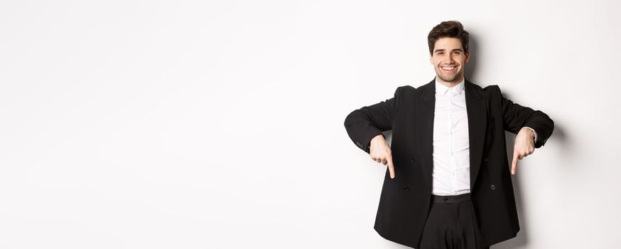 Portrait of good-looking stylish man in black suit, pointing fingers down and smiling, showing winter holidays promo, standing over white background.