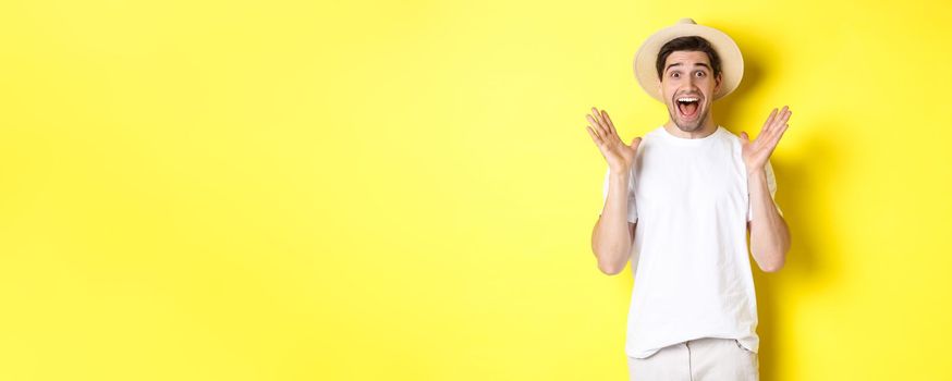 Concept of tourism and summer. Happy young man in straw hat looking amazed, reacting to surprise, standing over yellow background.