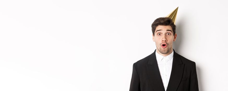 Close-up of attractive man in party hat and trendy suit, looking surprised, celebrating new year, standing against white background.