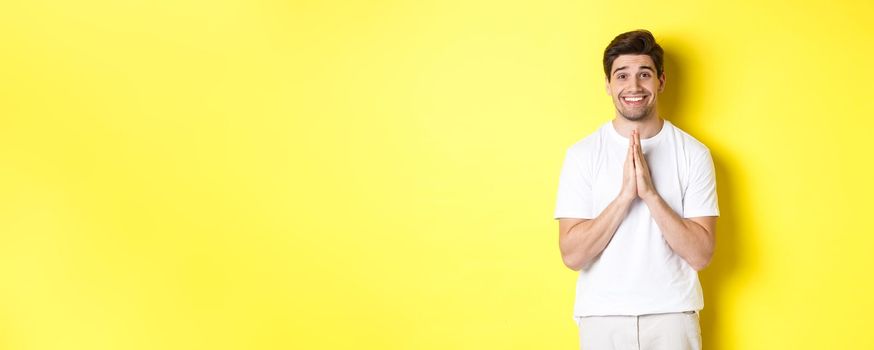 Hopeful man asking for help, begging favour, need something and smiling, standing over yellow background.