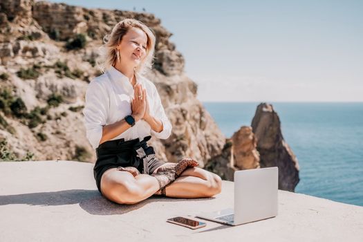 Happy girl doing yoga with laptop working at the beach. beautiful and calm business woman sitting with a laptop in a summer cafe in the lotus position meditating and relaxing. freelance girl remote work beach paradise