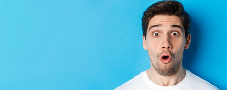 Headshot of surprised man gasping wondered, saying wow and looking amazed at camera, standing over blue background.