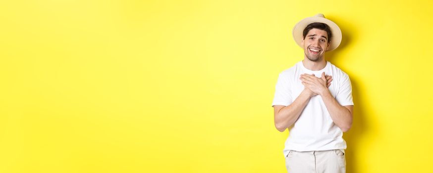 Concept of tourism and summer. Grateful guy in straw hat holding hands on heart, saying thank you and smiling with gratitude, standing against yellow background.