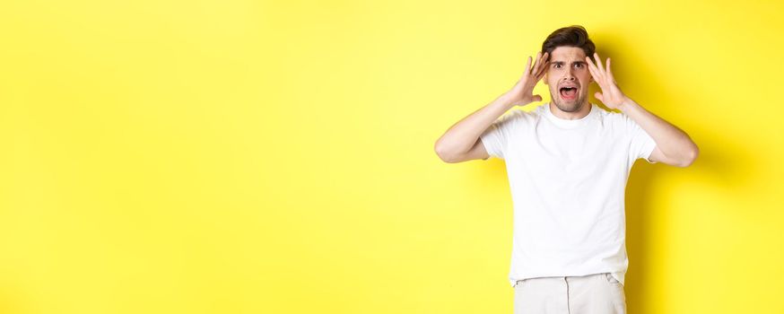 Frustrated guy looking alarmed, holding hands on head and feeling confused, panicking, standing over yellow background.