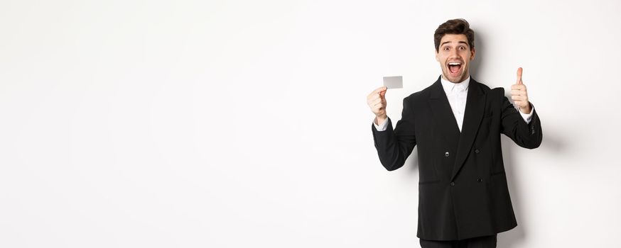 Image of good-looking businessman in black suit, showing thumbs-up and credit card, recommending bank, standing against white background.