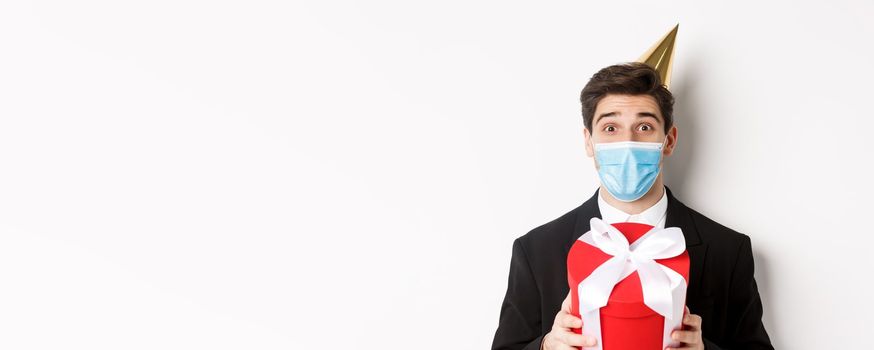 Concept of party during covid-19. Close-up of happy guy in trendy suit and medical mask, celebrating christmas, holding present and smiling, standing over white background.