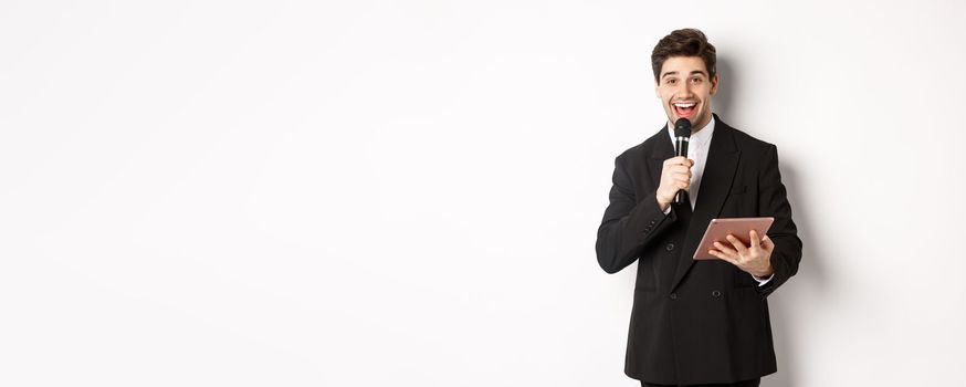 Image of handsome male performer, giving a speech, entertain people at party, holding microphone and digital tablet, standing over white background.