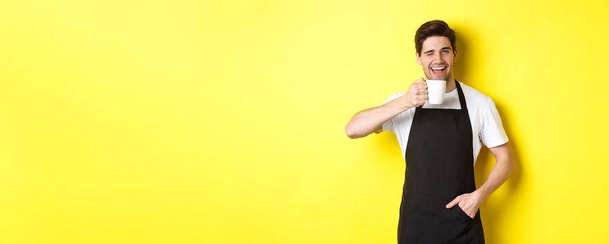 Handsome barista drinking cup of coffee and winking, inviting to cafe, standing over yellow background.