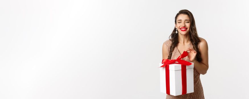 Merry Christmas. Charming woman in elegant dress open gift box and smiling delighted, unwrapping a present, standing over white background.