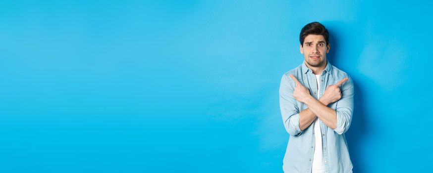 Portrait of nervous guy pointing fingers sideways, looking indecisive and asking for help with choice, showing left and right promo offers, standing against blue background.