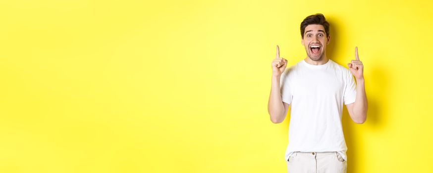 Portrait of excited handsome man in white t-shirt, pointing fingers up, showing offer, standing against yellow background.