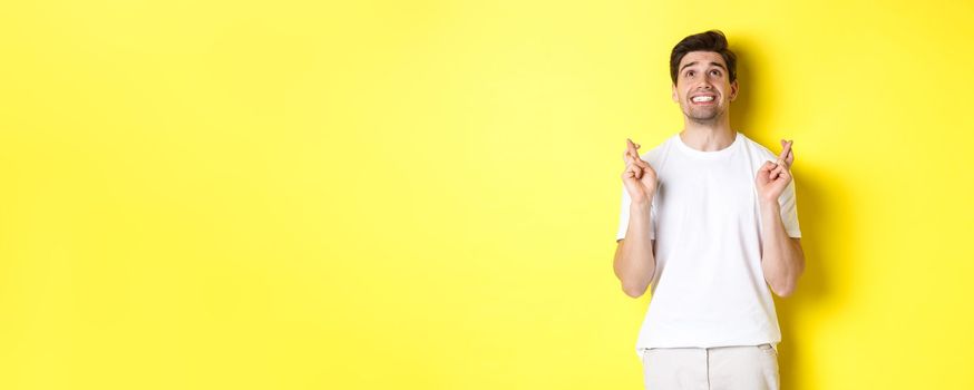 Nervous and hopeful man praying to god, making wish with fingers crossed, panicking and standing over yellow background.