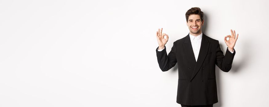 Concept of new year party, celebration and lifestyle. Portrait of handsome attractive man in black suit, smiling and showing okay signs, approve and recommend something, white background.