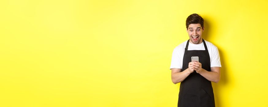 Barista looking surprised as reading message on mobile phone, standing in black apron against yellow background.