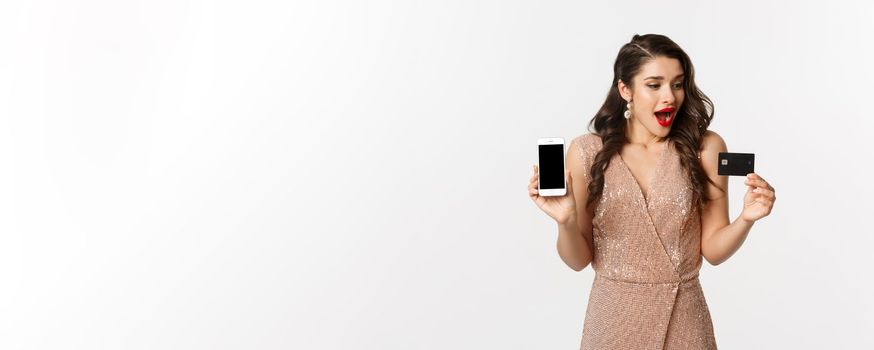 Online shopping and holidays concept. Attractive woman looking amazed at credit card and showing smartphone screen application, white background.