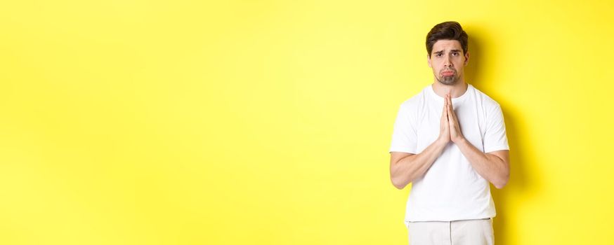 Sad guy begging for something, sulking and asking for favour, saying sorry, standing over yellow background.