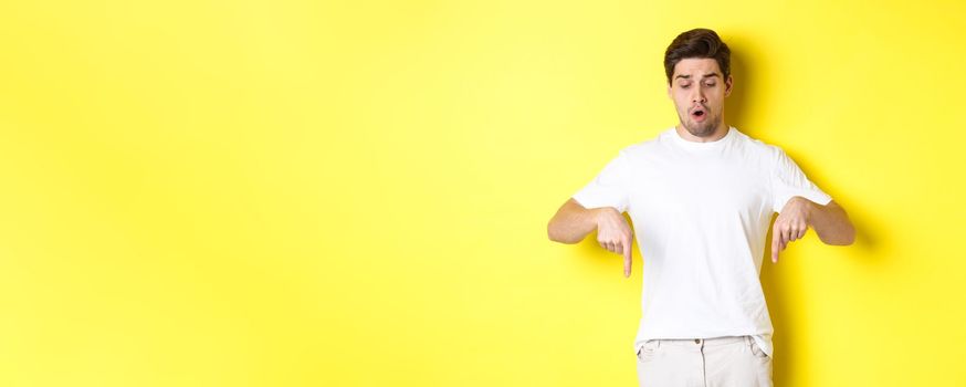 Worried guy pointing fingers and looking down, standing startled against yellow background.