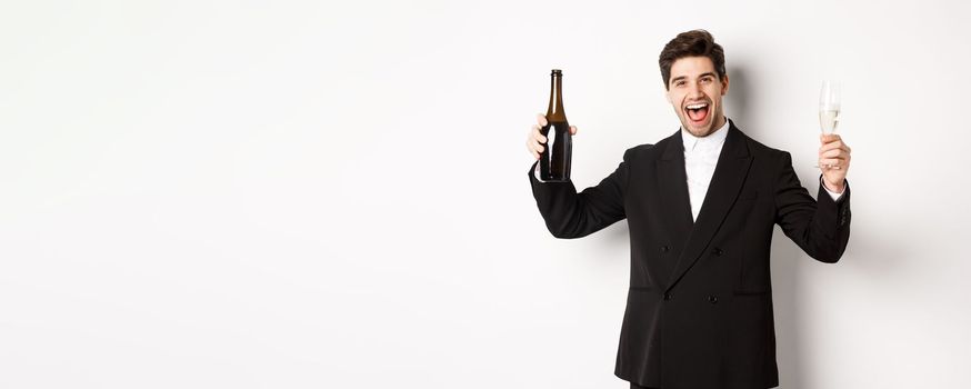 Concept of holidays, party and celebration. Handsome man in trendy suit having fun, holding bottle and glass of champagne, standing over white background.