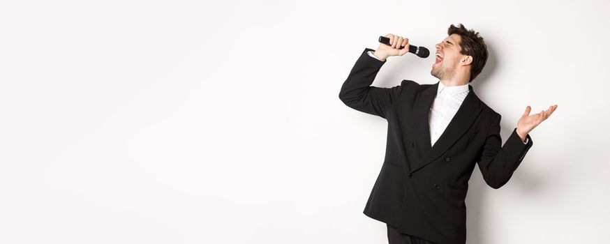 Portrait of handsome man singing a song with passion, standing in black suit, holding microphone and performning, posing over white background.