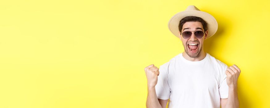 Concept of tourism and holidays. Lucky man tourist winning tickets, making fist pump and saying yes, wearing vacation clothes, standing over yellow background.