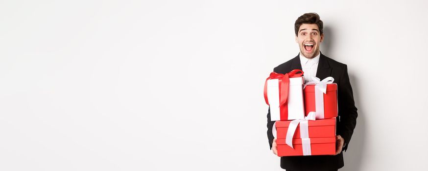 Concept of christmas holidays, celebration and lifestyle. Image of handsome amazed guy in suit, holding new year presents and smiling, standing against white background.