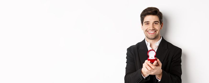 Image of handsome man looking romantic, open small box with engagement ring, making a proposal and smiling, standing against white background.