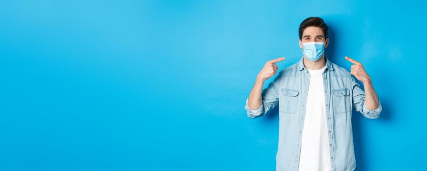 Concept of coronavirus, quarantine and social distancing. Handsome man pointing at medical mask and smiling, protection from virus spread during pandemic, blue background.