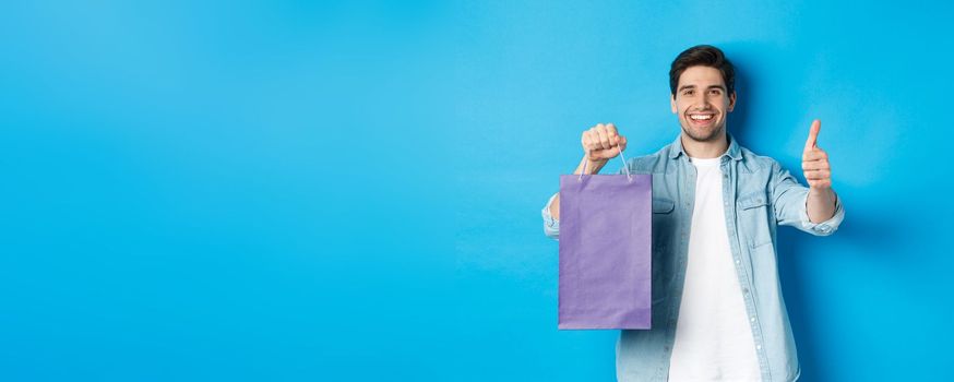 Concept of shopping, holidays and lifestyle. Satisfied smiling man holding paper bag, showing thumb-up and recommending store, standing over blue background.