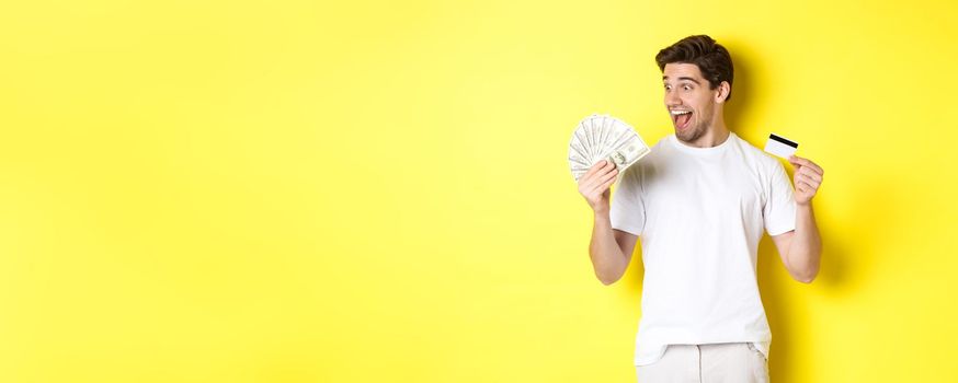 Cheerful guy looking at money, holding credit card, concept of bank credit and loans, standing over yellow background.