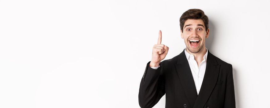 Close-up of handsome businessman in black suit, smiling amazed, showing number one, standing over white background.